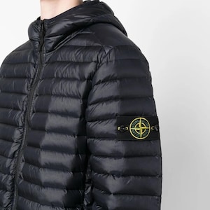 Könnte beinhalten: Eine schwarze, gesteppte Daunenjacke mit Kapuze und durchgehendem Frontreißverschluss. Am linken Ärmel befindet sich ein rechteckiger schwarzer Aufnäher mit einem gelb-grünen Kompass-Logo und dem Schriftzug Stone Island in Schwarz.