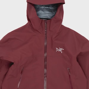 Könnte beinhalten: Eine dunkelrote Kapuzenjacke mit durchgehendem Frontreißverschluss, zwei seitlichen Reißverschlusstaschen und einer Kapuze. Der Markenname Arc'teryx ist auf der Kapuze aufgedruckt und das weiße Fossil-Logo ist auf der linken Brustseite eingestickt.