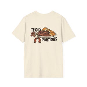 May include: A cream-coloured t-shirt with a graphic of a cowboy carrying a plate of food. The plate shows meat, beans, and cornbread, with the words "TEXAS PORTIONS" above and below the plate in a bold font.
