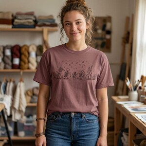Può includere: Una t-shirt a maniche corte color rosa polvere con una grafica a linee marrone scuro che raffigura un gruppo di insetti che suonano strumenti musicali, tra cui un violino, un tamburo e un contrabbasso, accompagnati da note musicali e stelle.