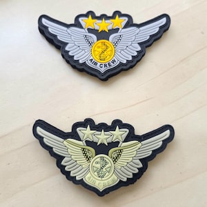 May include: Two air crew patches featuring a central circular emblem with an anchor, flanked by detailed wings and topped with three stars. The top patch has yellow stars and a yellow center, while the bottom patch features a muted, pale green color scheme.
