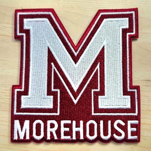 May include: A maroon and white embroidered patch featuring a large block letter M with the word MOREHOUSE printed in white capital letters underneath. The design has a thick maroon border and a white outline surrounding the letter and text.