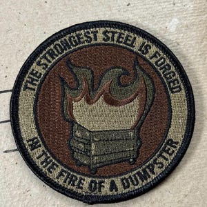 Puede incluir: Un parche bordado circular con un borde negro. El centro presenta un fondo marrón con un contenedor de basura en llamas. El texto "THE STRONGEST STEEL IS FORGED IN THE FIRE OF A DUMPSTER" está bordado en negro a lo largo del borde beige.