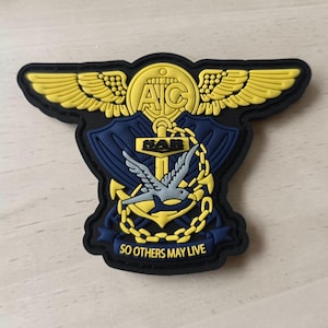 May include: A black, yellow, and grey rubberized patch featuring a central anchor, wings, and a bird. The design includes the text 'AIC' at the top, 'SAR' on the anchor, and the motto 'SO OTHERS MAY LIVE' on a banner at the bottom of the emblem.