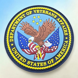 May include: A circular patch featuring the Department of Veterans Affairs seal. It displays a brown eagle with a white head, two American flags, and five yellow stars on a light blue background, all enclosed by a yellow rope-style border.