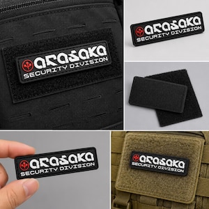 May include: A rectangular black tactical patch featuring the white text Arasaka Security Division and a red circular logo with a three-pronged symbol. The patch has a hook-and-loop backing for attachment to gear like bags or vests, shown in various settings.