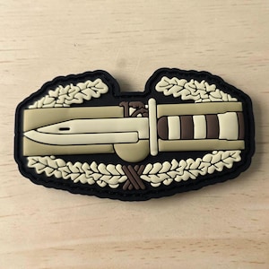 Puede incluir: Un parche rectangular con fondo negro y un diseño moldeado en color beige y marrón. El gráfico central muestra un cuchillo bayoneta horizontal centrado dentro de una corona de hojas de roble. El diseño presenta un relieve texturizado.
