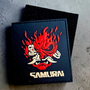 May include: A square black tactical patch featuring a raised, stylised red and gold graphic of a mechanical samurai mask surrounded by flames. The word SAMURAI is printed in a bold, blocky, gold-coloured font at the bottom of the patch.