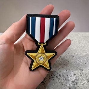 Pode incluir: Um emblema decorativo de medalha com uma estrela dourada de cinco pontas e um emblema circular central contendo uma pequena estrela branca. A medalha está suspensa por uma fita retangular com listras verticais nas cores azul-marinho, branco e vermelho.
