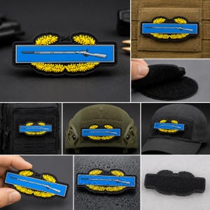 Può includere: Una toppa rettangolare con una barra orizzontale blu e l'illustrazione di un fucile bianco, incorniciata da una corona di foglie di quercia gialle su sfondo nero. La toppa è dotata di un retro in velcro per il fissaggio all'attrezzatura.