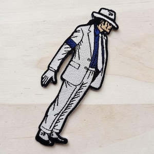 May include: An embroidered patch depicting a figure leaning forward at an angle, wearing a white suit, a blue shirt, a white fedora hat, and a single white glove. The design features black outlines and detailed stitching throughout the entire garment.