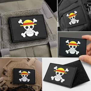 Puede incluir: Un parche rectangular negro con un diseño de calavera y tibias cruzadas en blanco, que lleva un sombrero de paja amarillo con una banda roja. El parche tiene un reverso de velcro y se muestra fijado en equipo táctico y sostenido a mano.