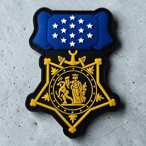 Pode incluir: Um emblema de borracha com o formato da Medalha de Honra. A seção superior é uma barra retangular azul com treze estrelas brancas. A seção inferior é uma estrela amarela de cinco pontas com um emblema circular central com duas figuras.