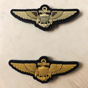 May include: Two military-style aviation insignia patches featuring a central shield and anchor design flanked by wings. The top patch is tan and black, while the bottom patch is gold and black. Both feature a raised, molded rubberized texture.