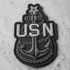 May include: A black and grey tactical patch featuring a central anchor design wrapped in a chain. The letters USN are prominently displayed in a bold, block font across the centre of the anchor, topped with a five-pointed star at the very peak.