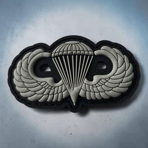 May include: A tactical morale patch featuring a raised, light gray rubberized design of a parachute centered between two stylized wings, all set against a solid black background with a contoured edge and a matte finish.