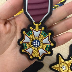 May include: A rubberized morale patch shaped like a military-style medal. The design features a maroon ribbon top section and a central circular emblem with a blue field containing seven white stars, surrounded by a white and gold cross-shaped frame.