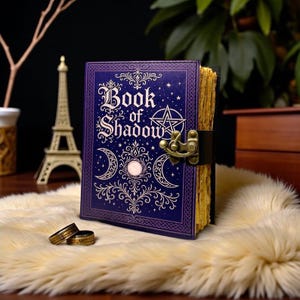 May include: A purple hardcover journal titled Book of Shadow with gold foil celestial designs including a pentagram and crescent moons. The book features a brass-colored metal latch closure and thick, textured, cream-colored pages with deckled edges.