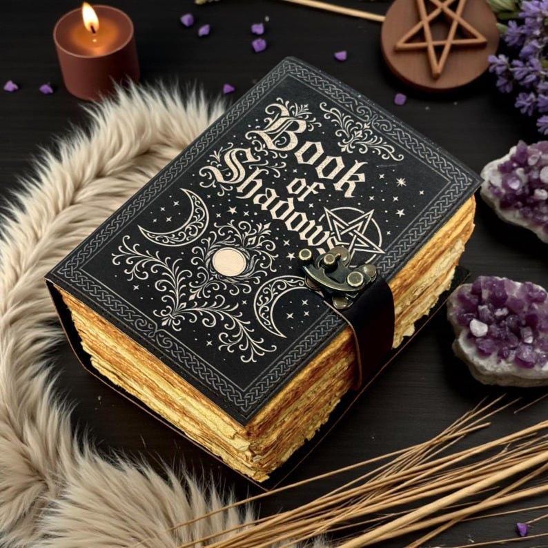 May include: A black hardcover book titled Book of Shadows with gold foil celestial designs including moons, stars, and a pentagram. The book features a metal latch closure and thick, deckled-edge pages that appear aged and tan in color.