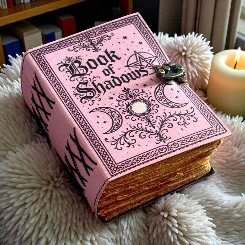 May include: A pink hardcover book titled Book of Shadows with black ink illustrations of a pentagram, crescent moons, and ornate scrollwork. The book features a metal latch closure and thick, textured, tan-colored pages with a deckled edge finish.