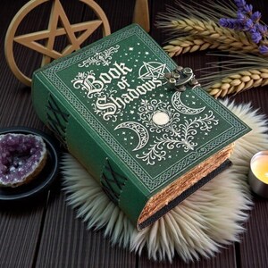 May include: A green hardcover book titled Book of Shadows with gold embossed celestial and pentagram designs on the front cover. The book features a metal latch closure, decorative side lacing, and textured pages with a weathered appearance.