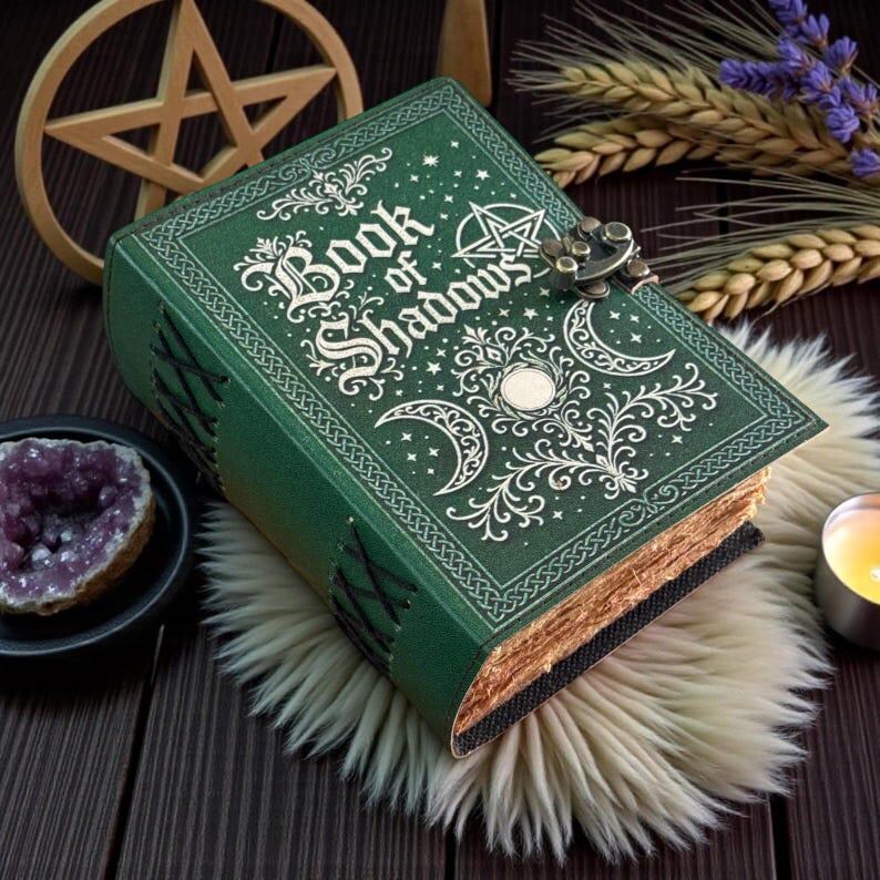 May include: A green hardcover book titled Book of Shadows with gold embossed celestial and pentagram designs on the front cover. The book features a metal latch closure, decorative side lacing, and textured pages with a weathered appearance.