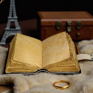 May include: An open journal with thick, textured, cream-colored pages and a dark binding. A simple gold-toned metal ring rests on the soft, light-colored surface in front of the book, with a miniature Eiffel Tower model and a brown box nearby.