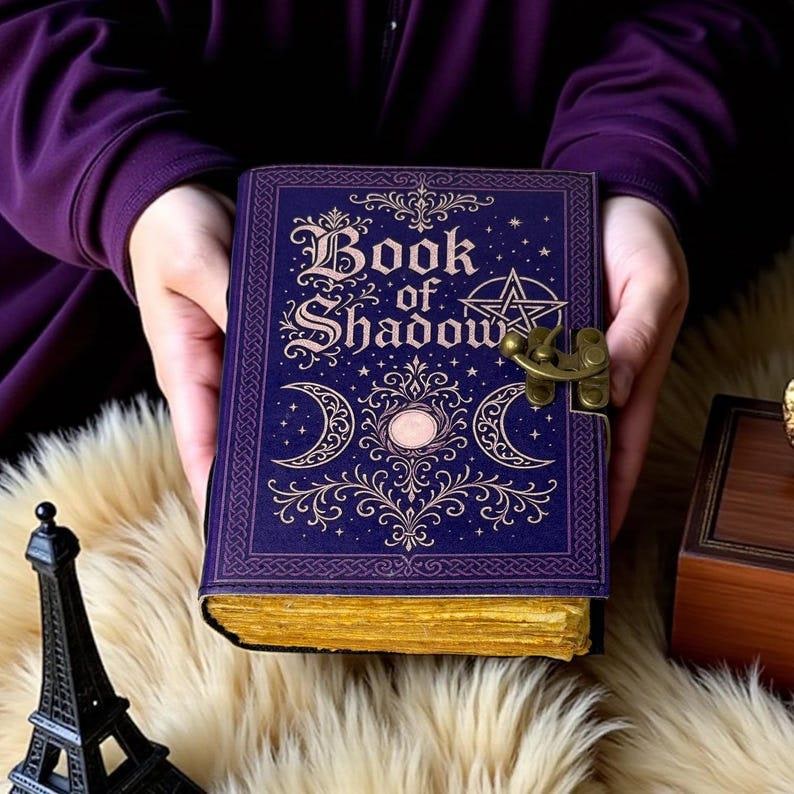 May include: A rectangular purple journal with the text Book of Shadow printed in gold on the front cover. The cover features gold foil illustrations of a pentagram, crescent moons, and ornate scrollwork. It has a brass latch and gold-edged pages.