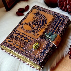 Könnte beinhalten: Ein rechteckiges Lederjournal mit einem geprägten Drachenmotiv auf dem Vorderdeckel. Der Einband hat dunkelbraune Zierränder, einen Metallverschluss und eine mittige, ovale Edelsteineinlage. Die Seiten haben goldfarbene Kanten.