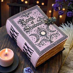 May include: A light pink journal titled Book of Shadows with black ink illustrations of moons, stars, and a pentagram. The book features a metal latch closure and a side binding with black cord lacing. The pages have a weathered, aged appearance.