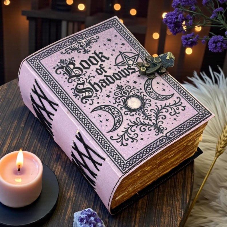May include: A light pink journal titled Book of Shadows with black ink illustrations of moons, stars, and a pentagram. The book features a metal latch closure and a side binding with black cord lacing. The pages have a weathered, aged appearance.