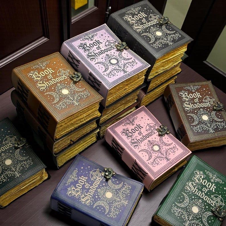 May include: A collection of rectangular journals titled Book of Shadows, featuring embossed celestial motifs, crescent moons, and pentagrams. Each book has a metal latch closure and deckled, gold-toned pages, available in black, brown, pink, purple, and green.