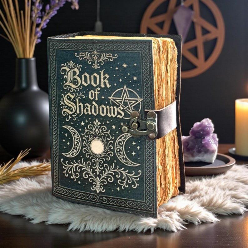 May include: A dark green leather-bound journal titled Book of Shadows with gold embossed celestial motifs including a pentagram and crescent moons. The book features a metal latch closure and thick, deckled-edge paper pages that appear aged.