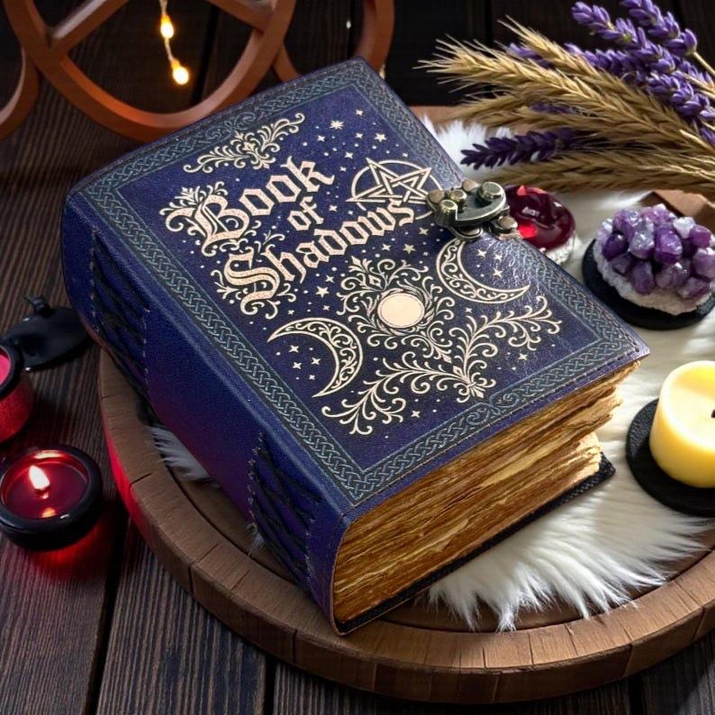 May include: A dark blue hardcover book titled Book of Shadows with gold embossed celestial designs including moons, stars, and a pentagram. The book features a metal latch closure and thick, deckled-edge pages with a visible stitched binding.