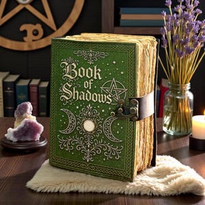 May include: A green hardcover book titled Book of Shadows with gold embossed celestial symbols, including a pentagram and crescent moons. The book features a dark brown leather strap with a metal latch and thick, textured, cream-colored pages.