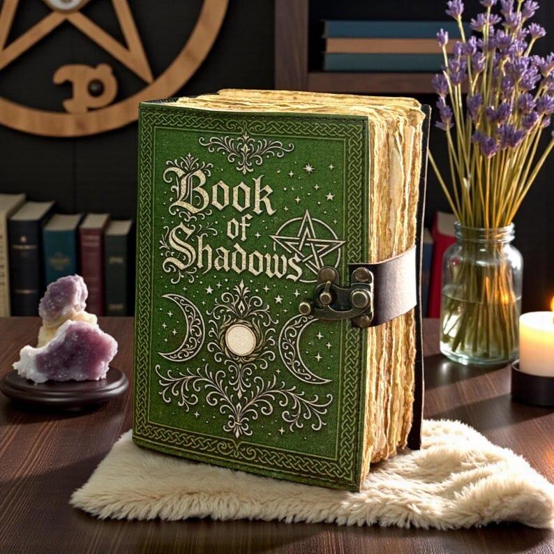 May include: A green hardcover book titled Book of Shadows with gold embossed celestial symbols, including a pentagram and crescent moons. The book features a dark brown leather strap with a metal latch and thick, textured, cream-colored pages.