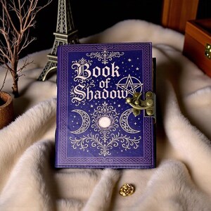 May include: A purple hardcover journal titled Book of Shadow with gold foil lettering, a pentagram, and moon phase illustrations. The book features an ornate border and a metallic brass-colored latch closure on the right side to keep it securely shut.