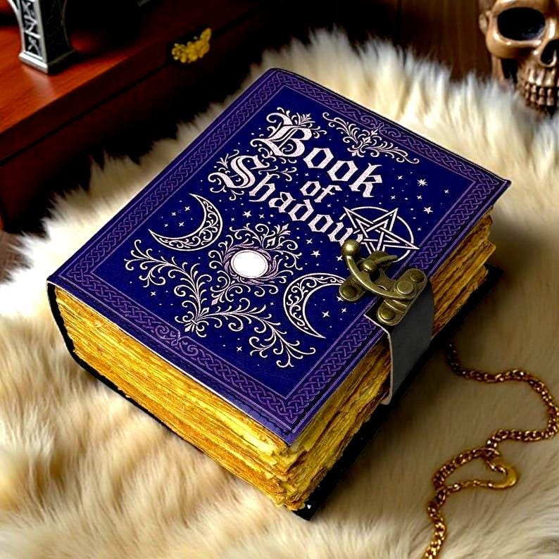 May include: A rectangular book titled Book of Shadows with a deep blue cover featuring white celestial motifs, including a crescent moon and a pentagram. The book has a brass-colored metal latch and thick, deckled-edge pages with a gold-toned finish.