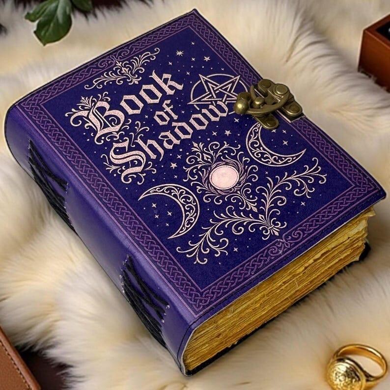 May include: A purple journal with the text Book of Shadow embossed in gold on the cover. The cover features gold celestial designs including a pentagram, crescent moons, and stars. A brass-colored metal latch secures the book, which has gold-edged pages.