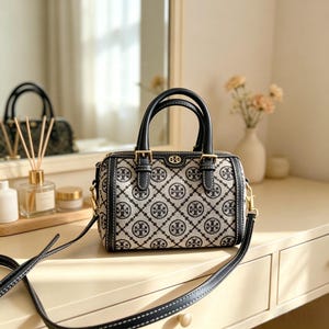 May include: A small handbag with a repeating logo pattern in black and cream, featuring black leather handles and a detachable shoulder strap. The bag has gold-coloured hardware. It is displayed on a light-coloured surface.
