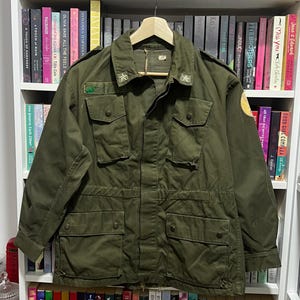 May include: An olive green military-style field jacket featuring a button-down front, four large front pockets with buttoned flaps, shoulder epaulets, and star insignias on the collar points. The jacket includes a circular patch on the sleeve.