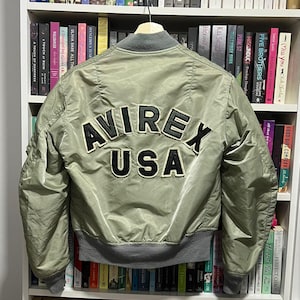 May include: An olive green bomber jacket featuring a grey ribbed collar, cuffs, and hem. The back displays large, black, embroidered lettering that reads AVIREX USA with a white border. The jacket is constructed from a smooth, satin-like material.