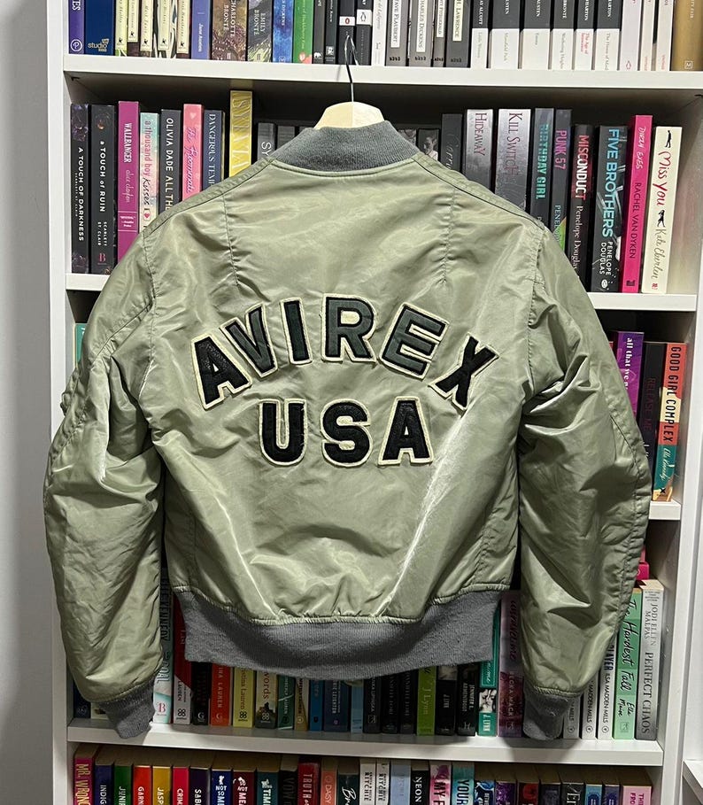 May include: An olive green bomber jacket featuring a grey ribbed collar, cuffs, and hem. The back displays large, black, embroidered lettering that reads AVIREX USA with a white border. The jacket is constructed from a smooth, satin-like material.