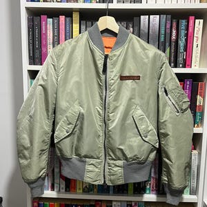May include: A sage green bomber jacket featuring a full-length front zipper, a ribbed gray collar, cuffs, and hem. The jacket includes two front snap-button pockets, a zippered utility pocket on the left sleeve, and a small brown rectangular patch.