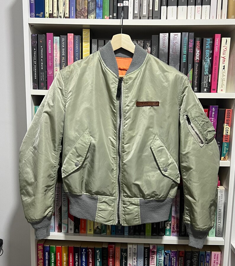 May include: A sage green bomber jacket featuring a full-length front zipper, a ribbed gray collar, cuffs, and hem. The jacket includes two front snap-button pockets, a zippered utility pocket on the left sleeve, and a small brown rectangular patch.