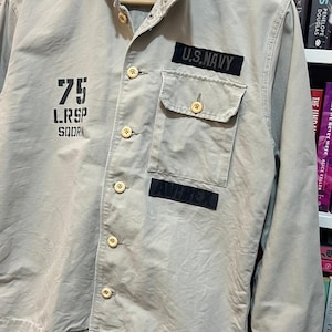 May include: A tan military-style jacket with button closures and two front pockets. The jacket features patches that read "U.S. NAVY" and "AVIREX", along with the number "75" and other text. The jacket is hanging on a wooden hanger.