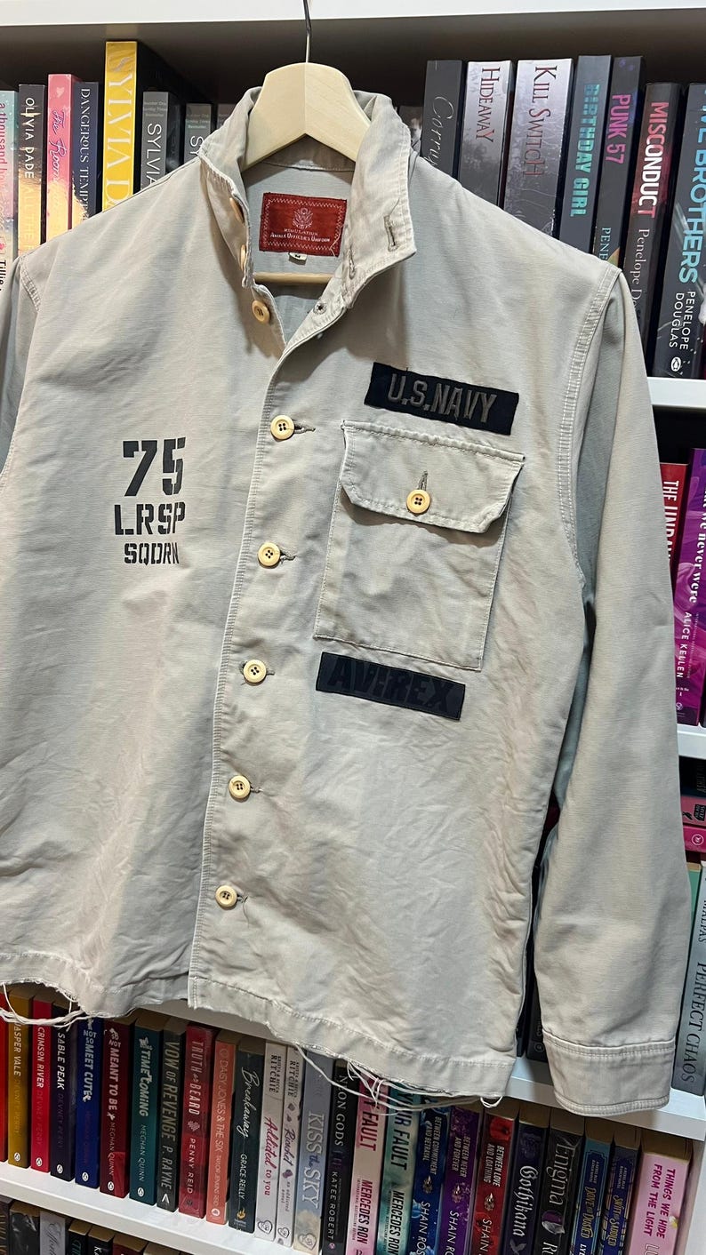 May include: A tan military-style jacket with button closures and two front pockets. The jacket features patches that read "U.S. NAVY" and "AVIREX", along with the number "75" and other text. The jacket is hanging on a wooden hanger.