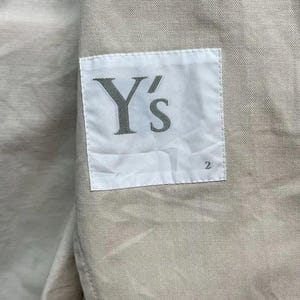 Puede incluir: Una prenda de tela beige con una etiqueta rectangular blanca cosida en la superficie. La etiqueta muestra el texto Y's en una fuente serif gris oscuro, con el n&uacute;mero 2 impreso en un tama&ntilde;o de fuente m&aacute;s peque&ntilde;o en la esquina inferior derecha.