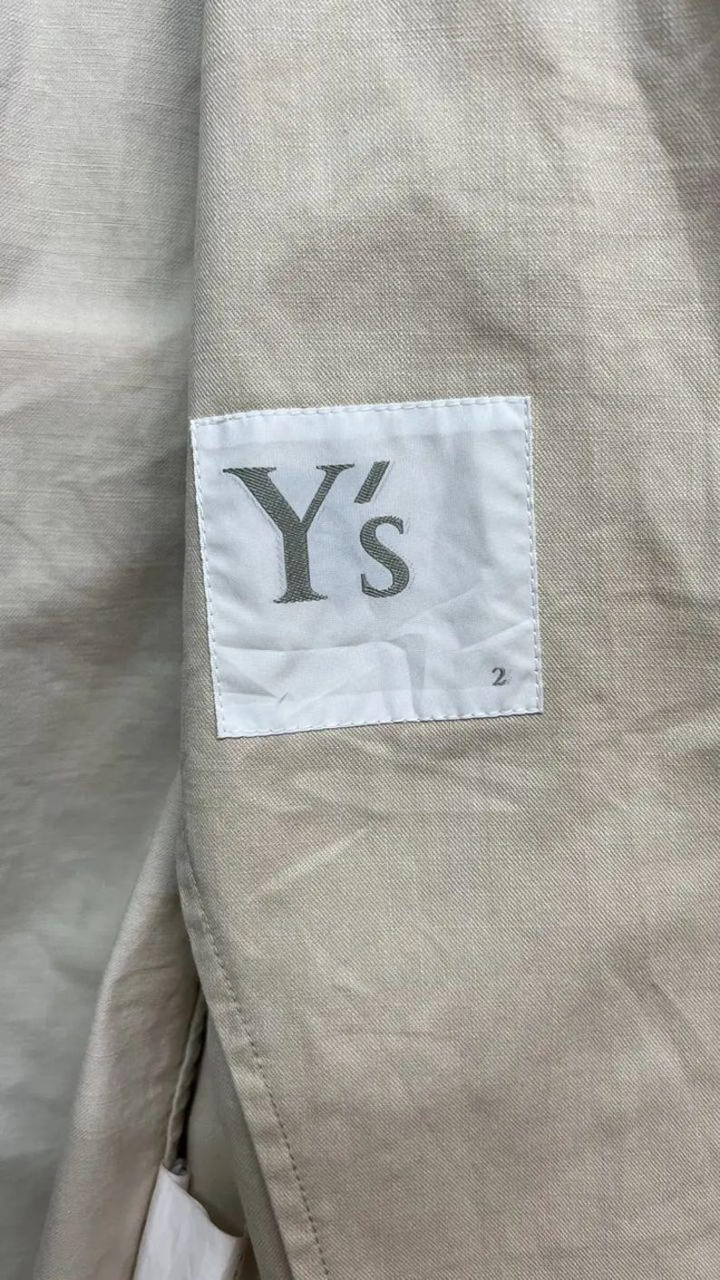 Puede incluir: Una prenda de tela beige con una etiqueta rectangular blanca cosida en la superficie. La etiqueta muestra el texto Y's en una fuente serif gris oscuro, con el n&uacute;mero 2 impreso en un tama&ntilde;o de fuente m&aacute;s peque&ntilde;o en la esquina inferior derecha.