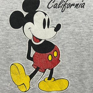 May include: A light grey hooded sweatshirt featuring a graphic of Mickey Mouse in black, white, red, and yellow. The character is positioned below the word California in black script. The Disney copyright symbol is printed below the character.